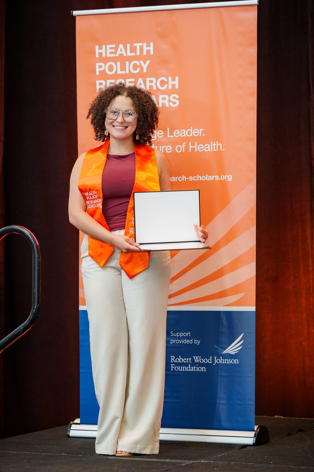 Scholar in an orange Health Policy Research Scholars stole holding a certificate at the completion ceremony
