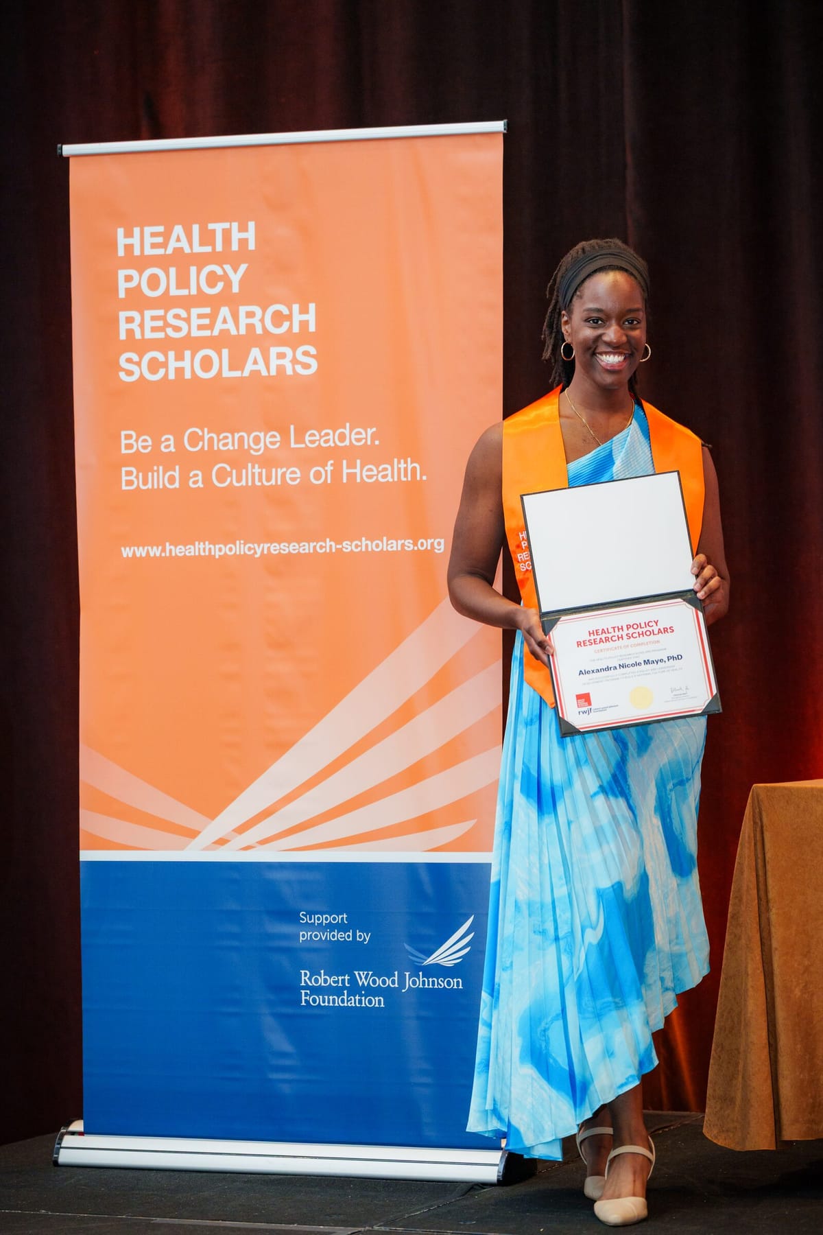 Graduating scholar holding a certificate beside the Health Policy Research Scholars banner