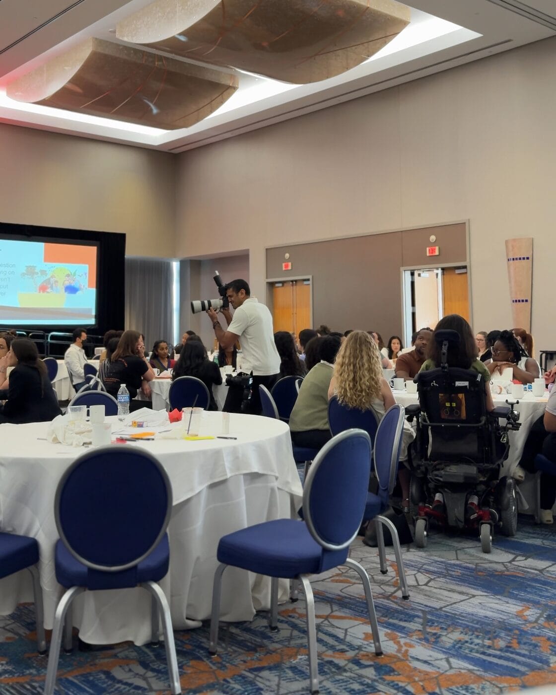 Simo photographing a session with a telephoto lens among the tables at the Cohort 9 convening in Baltimore